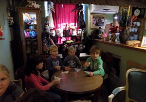 Czterech chłopców i dziewczynka siedzą przy okrągłym drewnianym stole z ciasteczkami w rękach. W tle rekwizyty teatralne.